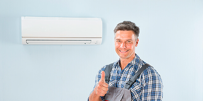Smiling technician in plaid shirt and overalls giving thumbs up near a mounted air conditioning unit.