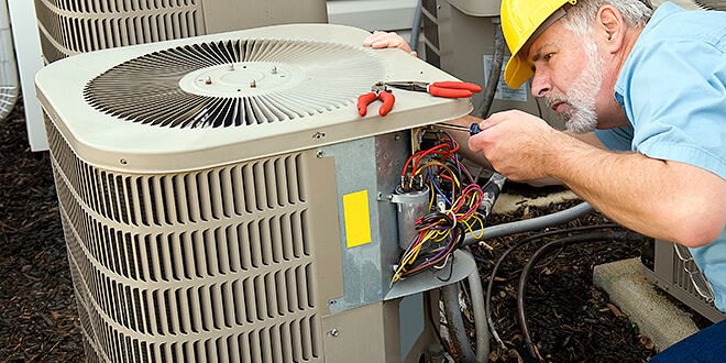 Technician in a yellow hard hat fixing an outdoor air conditioning unit with tools.