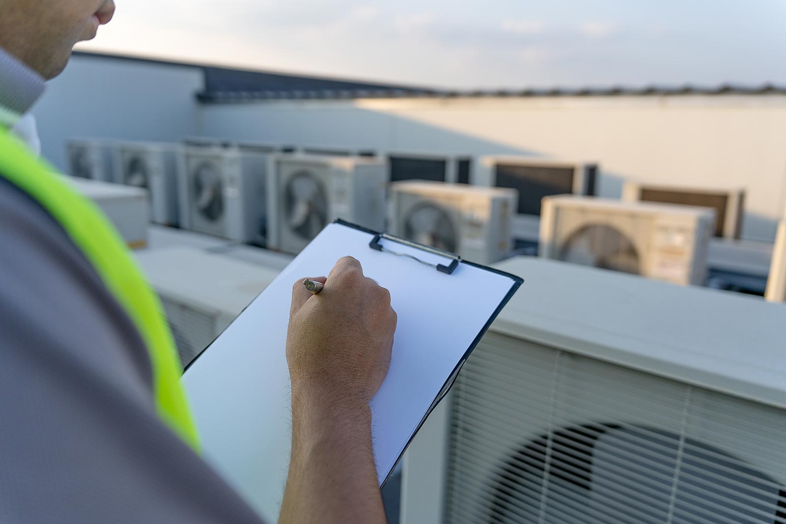 Technician in a high-visibility vest holding a clipboard, inspecting rooftop air conditioning units.