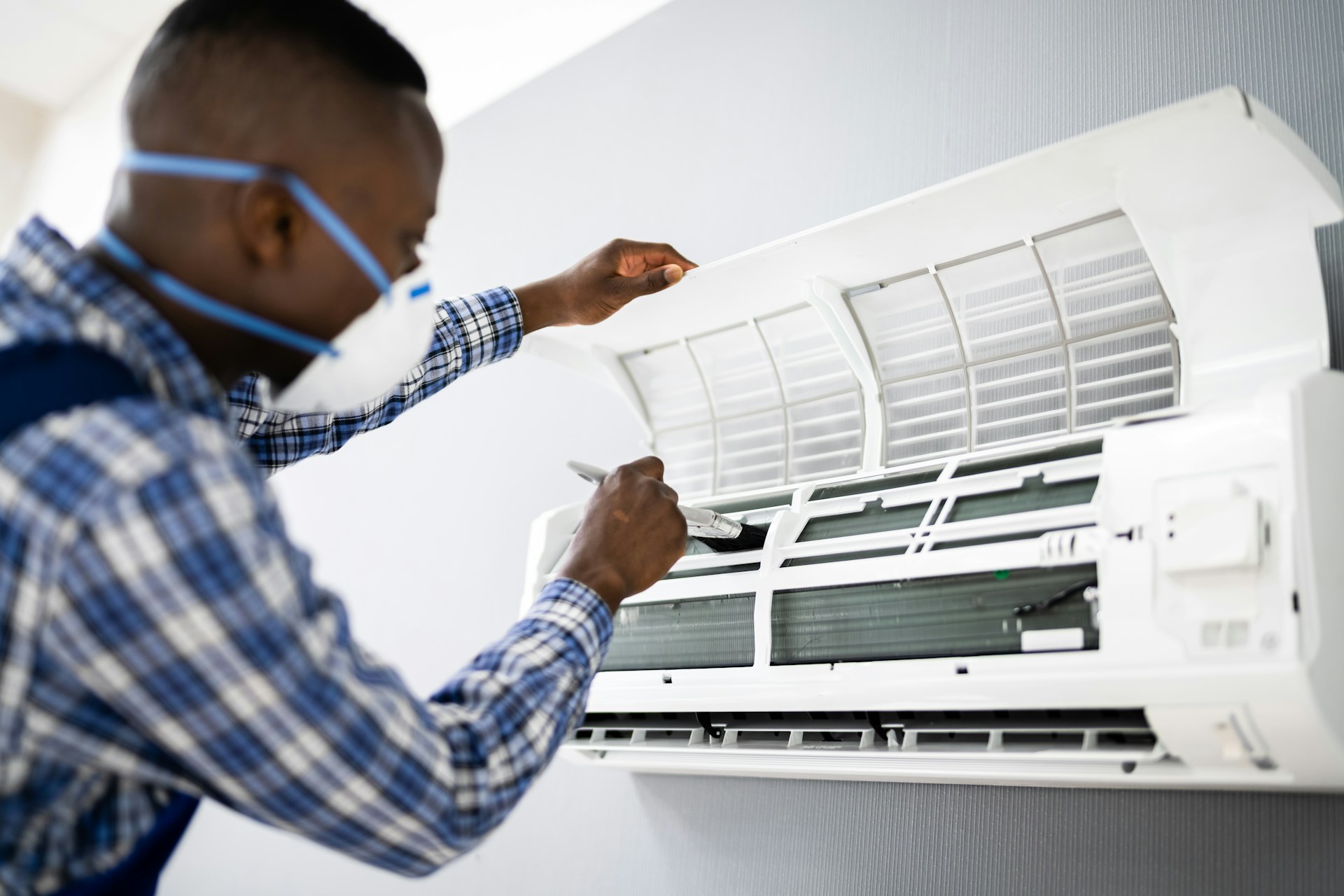 Technician wearing a mask cleans the interior of a wall-mounted air conditioner with a brush.