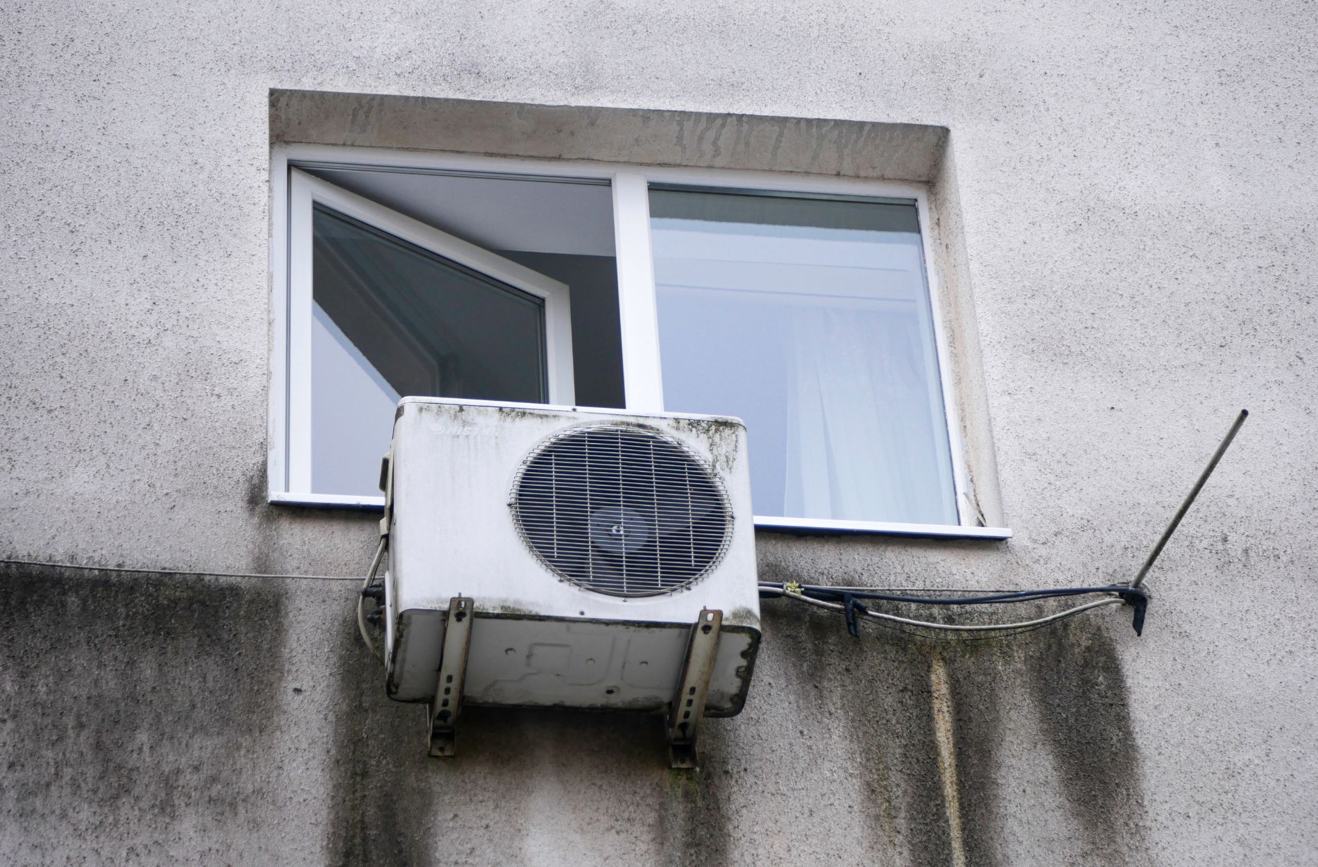 Wall-mounted air conditioning unit below an open window on a concrete building facade.