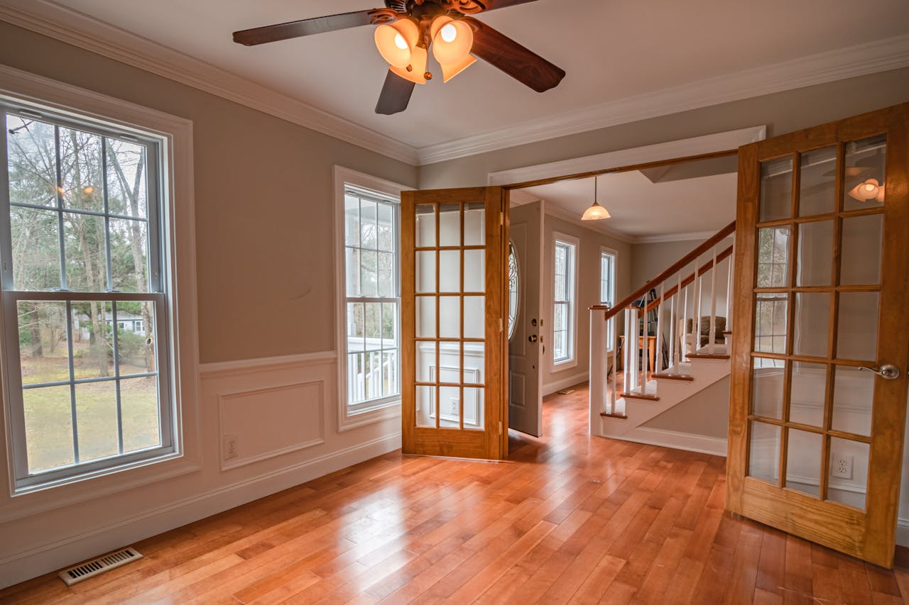 Bright room with hardwood floors, ceiling fan, glass-panel doors, large windows, and a staircase in the background.
