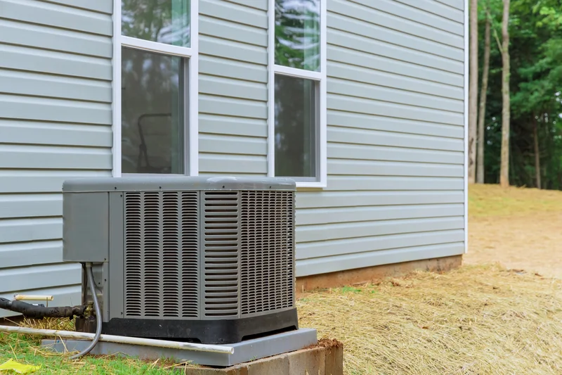 Exterior air conditioning unit next to a house with grey siding and two windows, set on a concrete pad in a grassy area.