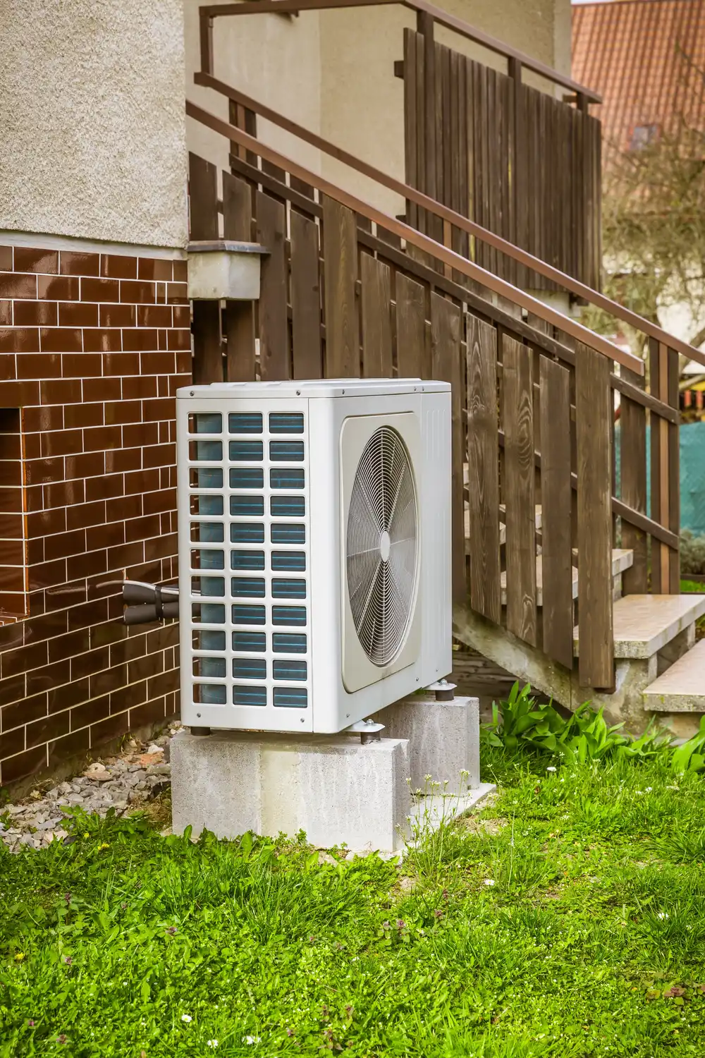 Exterior HVAC unit on concrete blocks beside a brick wall and wooden stairs, surrounded by green grass.