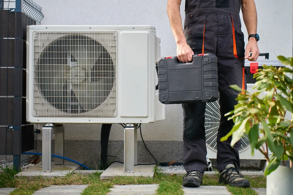 Technician holding a toolbox next to an outdoor air conditioning unit.