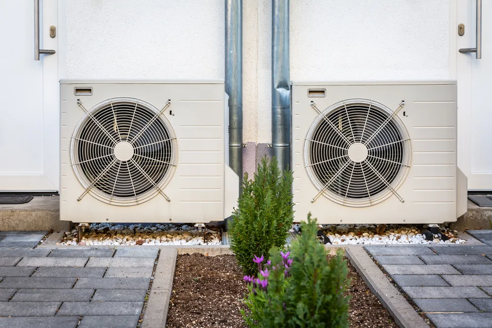 Two outdoor air conditioning units on a concrete patio, surrounded by small plants and paved stones.