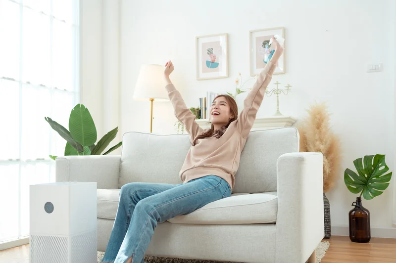 Woman relaxing on a sofa with arms raised, next to an air purifier, in a bright and cozy living room.