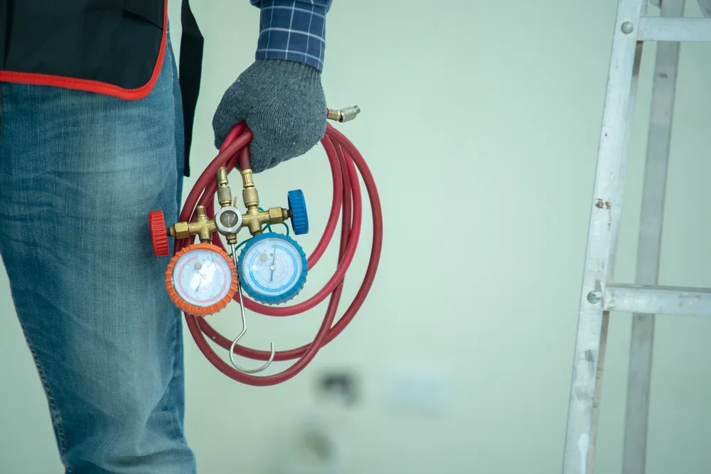 Technician in gloves holding HVAC manifold gauges next to a ladder.