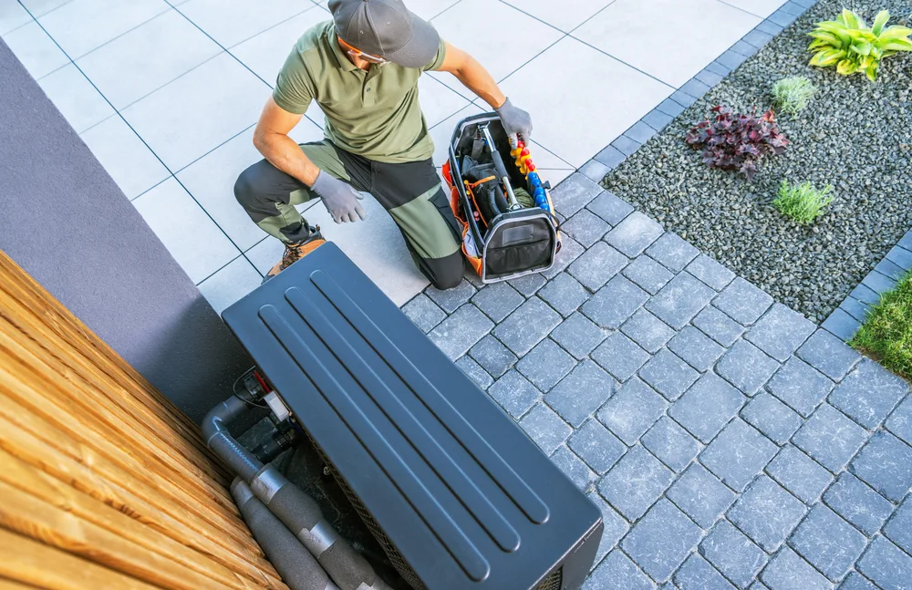 Technician kneeling outdoors beside an HVAC unit with an open tool bag on a paved patio.