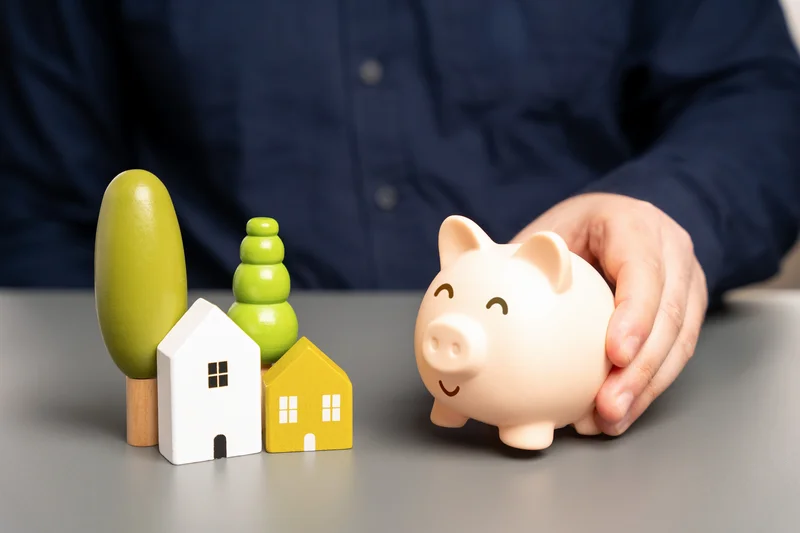 Hand holding a piggy bank next to wooden trees and house models on a table.