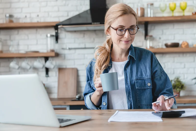 Woman holding a mug while working on papers at a kitchen counter with a laptop open.