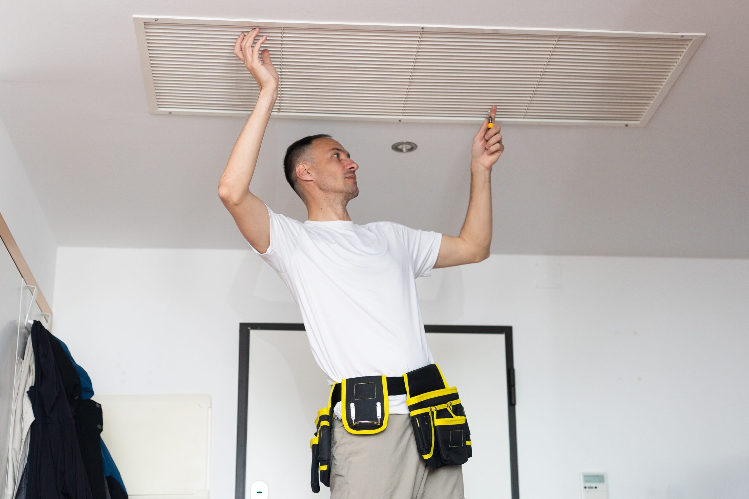 Technician adjusting ceiling vent with a screwdriver, wearing a tool belt.